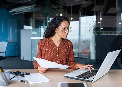 Young female professional seated at her desk, looking down at her laptop while holding sheets of paper in the other hand